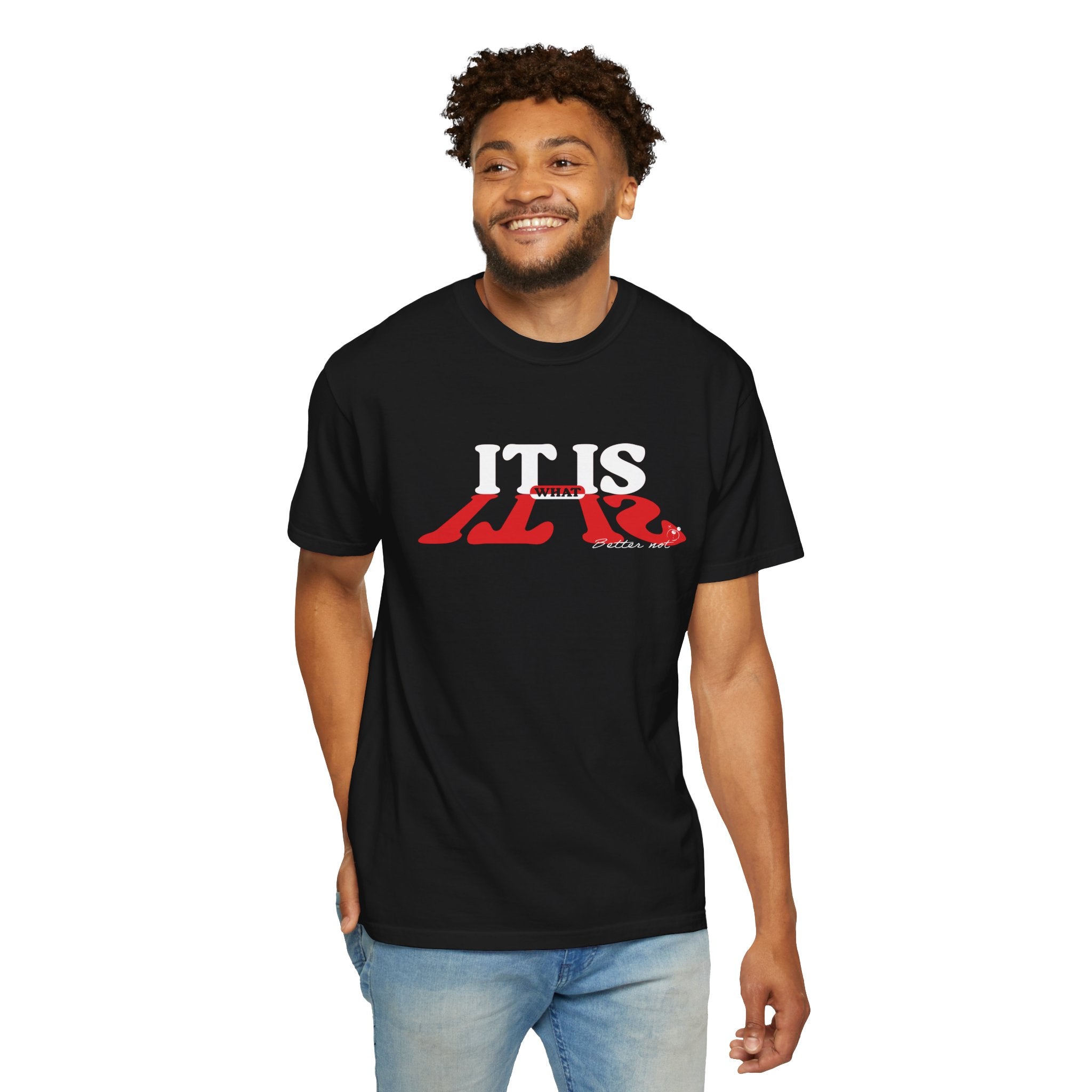 A curly-haired man is wearing the "It Is What It Is Tee" by Printify. The black T-shirt, made from 100% US cotton, features bold red and white text and complements his blue jeans as he smiles warmly.