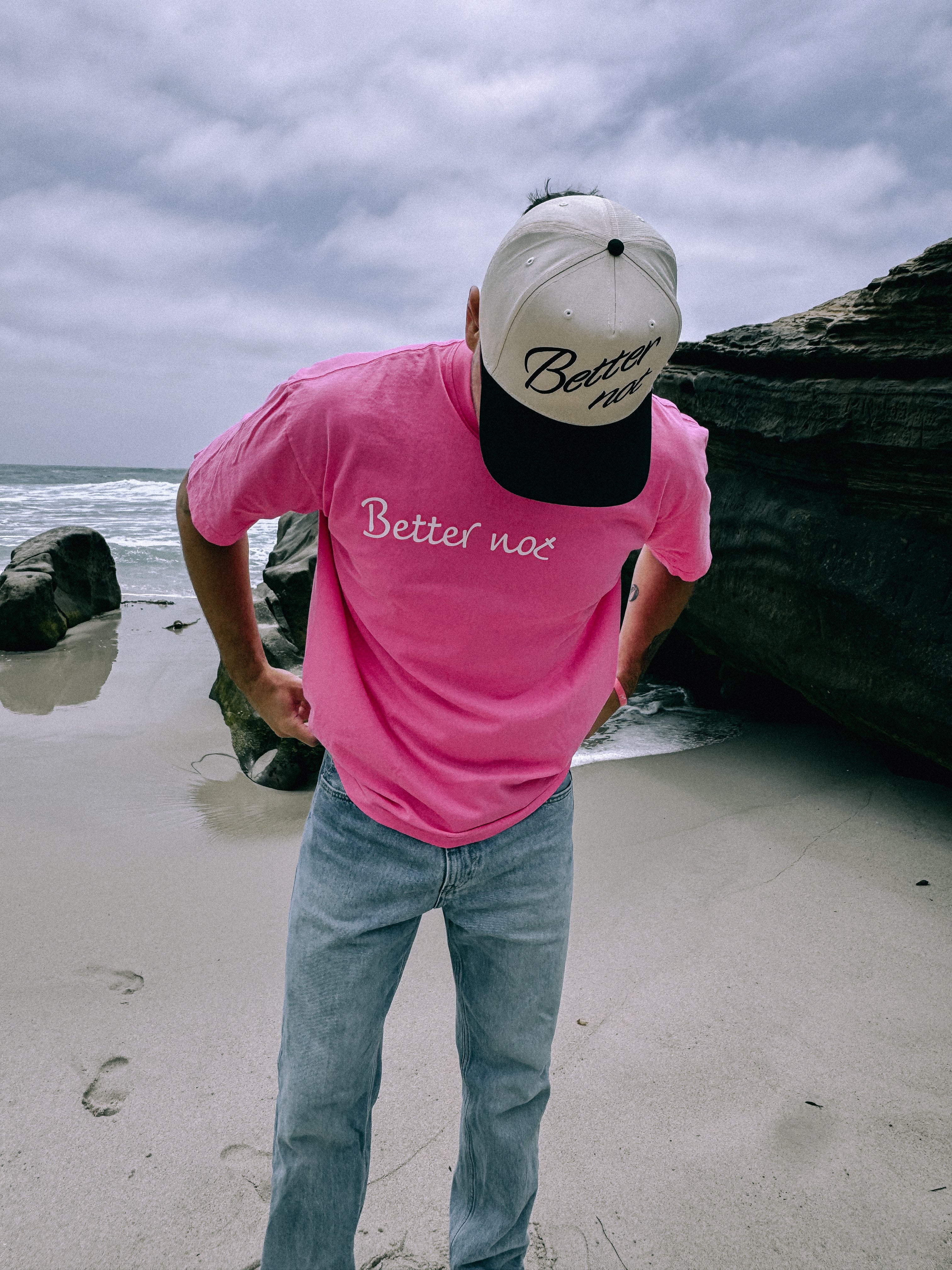 Wearing a "Better not" oversized Statement Pink Tee and beige cap, a person stands on a rocky beach, looking down. The unisex cotton apparel adds a carefree vibe to the serene coastal scene.