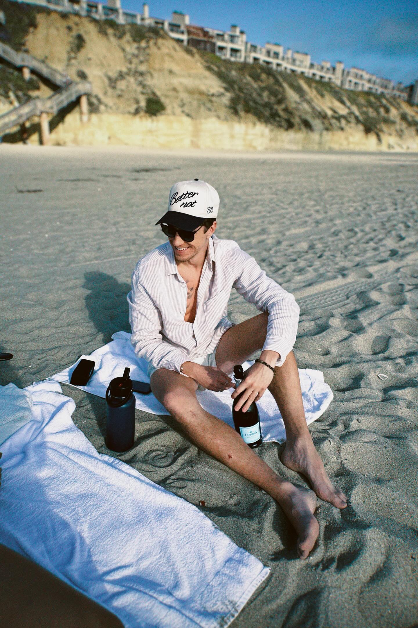 Person sitting on a sandy beach wearing a light cream cap with 3D embroidered artwork, sunglasses, and a long-sleeved shirt, holding The Classic by Better Not with a towel and other items around them. Bluff with houses in the background.