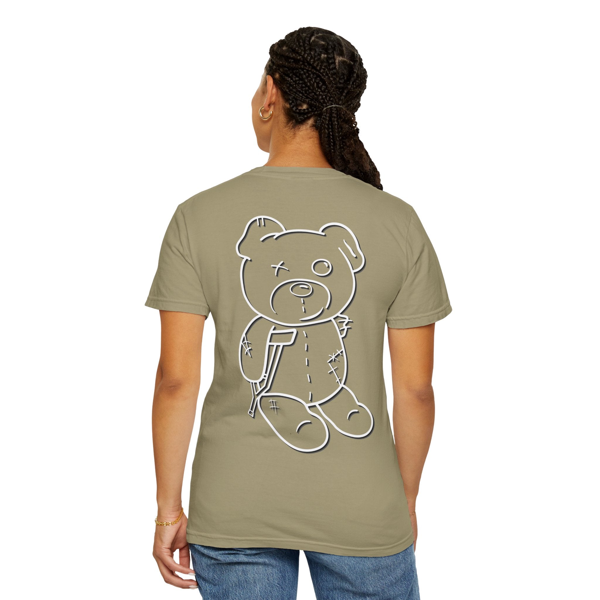 A person faces away wearing a khaki Printify Mascot Tee with a large cartoon teddy bear design on the back, paired with jeans and braided hair. Made from US cotton, this Comfort Colors premium tee blends style and comfort for any casual outing.