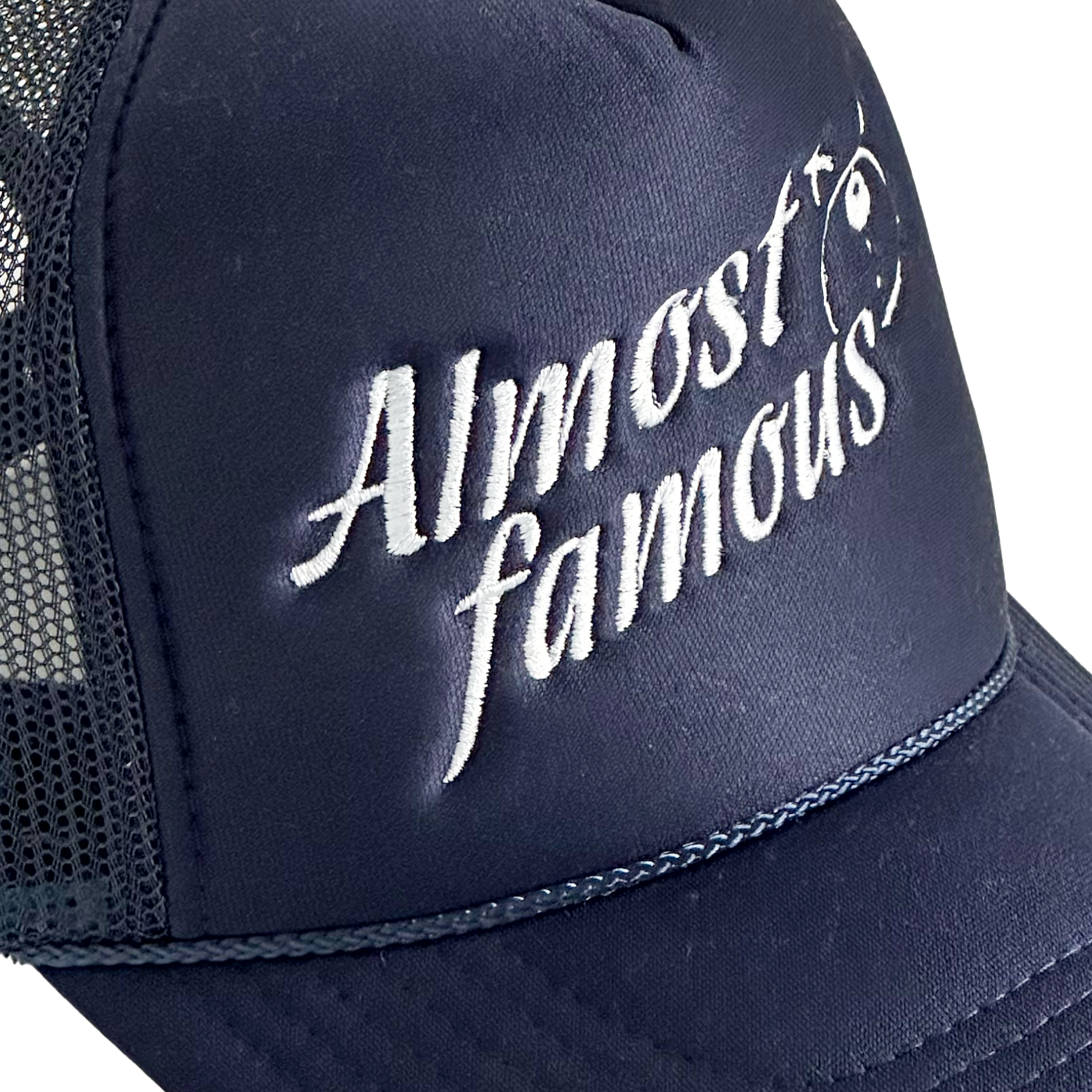 A close-up of the Almost Famous Trucker cap by Better not, featuring white "Almost Famous" embroidery on its dark blue front. This High-Crown Fit cap includes a mesh back and snapback closure.