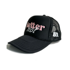 A black "Classic Trucker" cap from Better not, featuring white and pink "better not" embroidery on the front with a Mid Crown fit. The side of the cap displays the initials "BN" in white. It is pictured next to a product tag and is available for next day shipping.