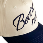 Close-up of The Classic baseball cap in beige and dark blue by Better not, with "Better now" embroidered in navy blue script on the front, featuring a snapback closure for adjustable comfort.