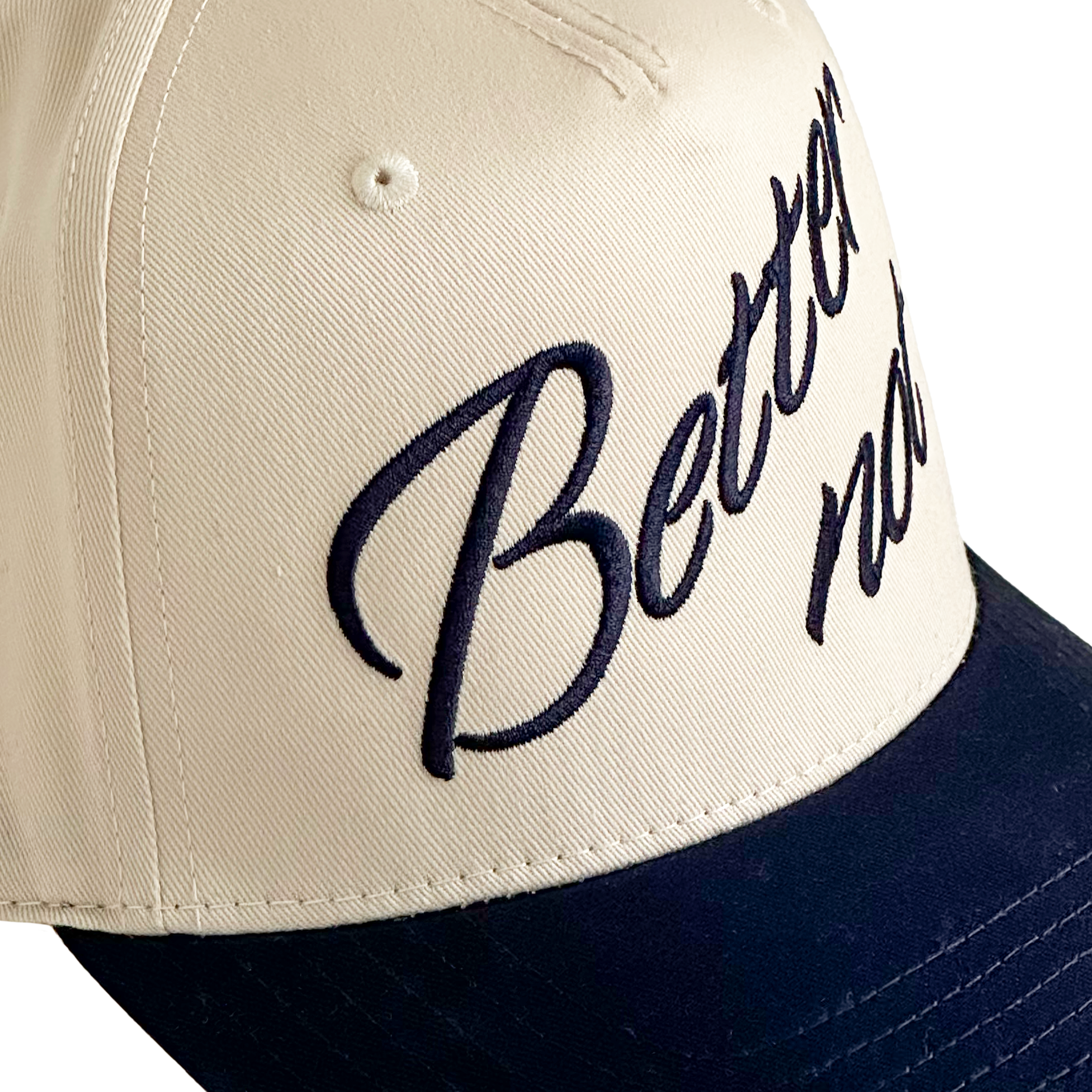 Close-up of The Classic baseball cap in beige and dark blue by Better not, with "Better now" embroidered in navy blue script on the front, featuring a snapback closure for adjustable comfort.