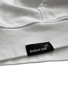 Close-up of the white Have Fun Be "Safe" Hoodie v2, crafted from brushed fleece, with a black label that has "Better not" and a small graphic symbol on it.