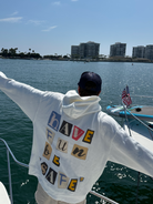 Person standing on a boat with arms outstretched, facing away from the camera, wearing the Better not "Have Fun Be Safe" Hoodie v2. Waterfront cityscape and clear sky in the background.