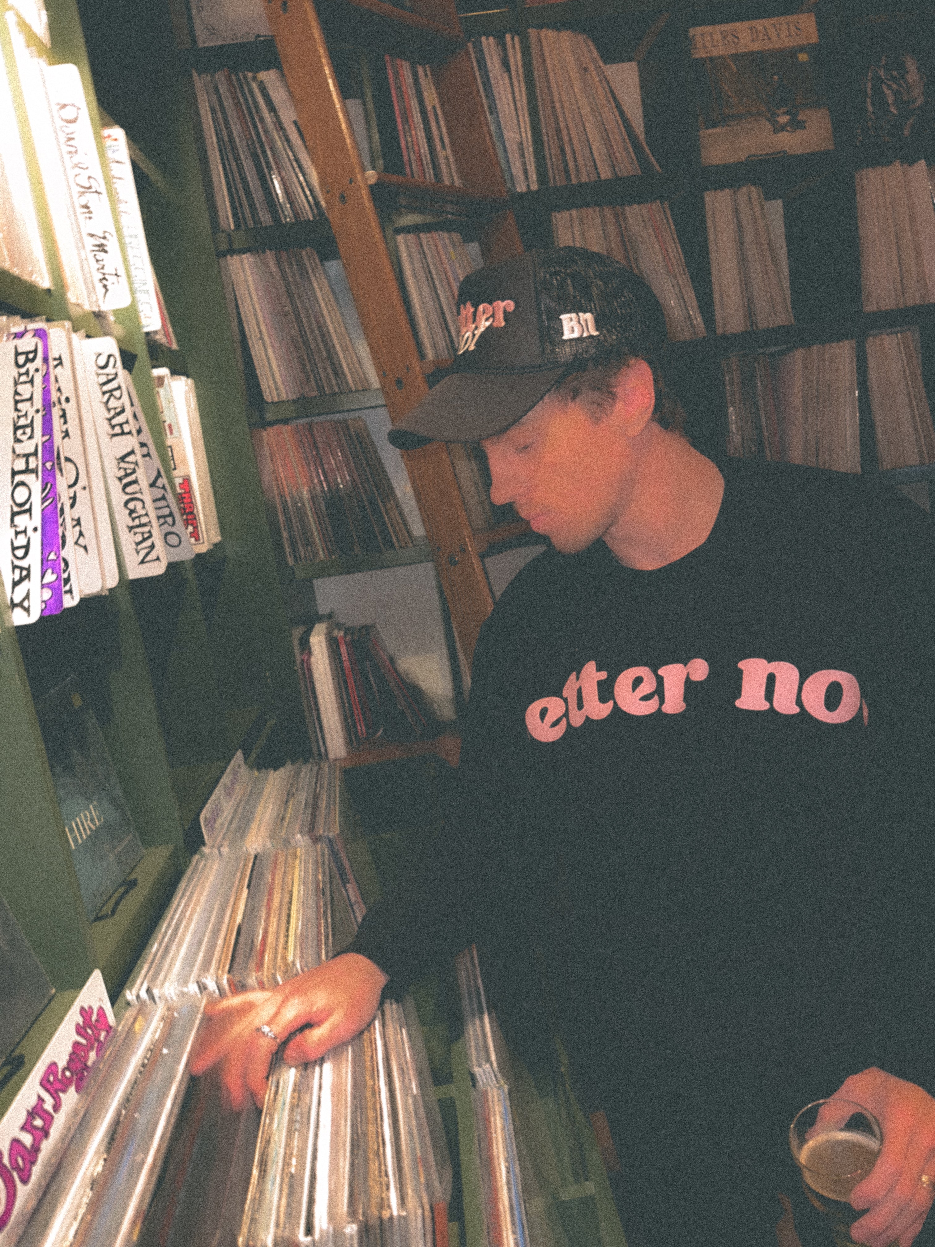 A young man in a black "Better not" Staple Crew sweatshirt with premium ring-spun cotton and cap browsing vinyl records in a dimly lit store, holding a coffee cup.