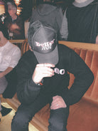 Description: Person wearing a black embroidered Better not Classic Trucker (mid crown) hat and a black sweatshirt with indistinguishable white text, sitting on an orange sofa with people in the background. The person is resting their hand on their knee.