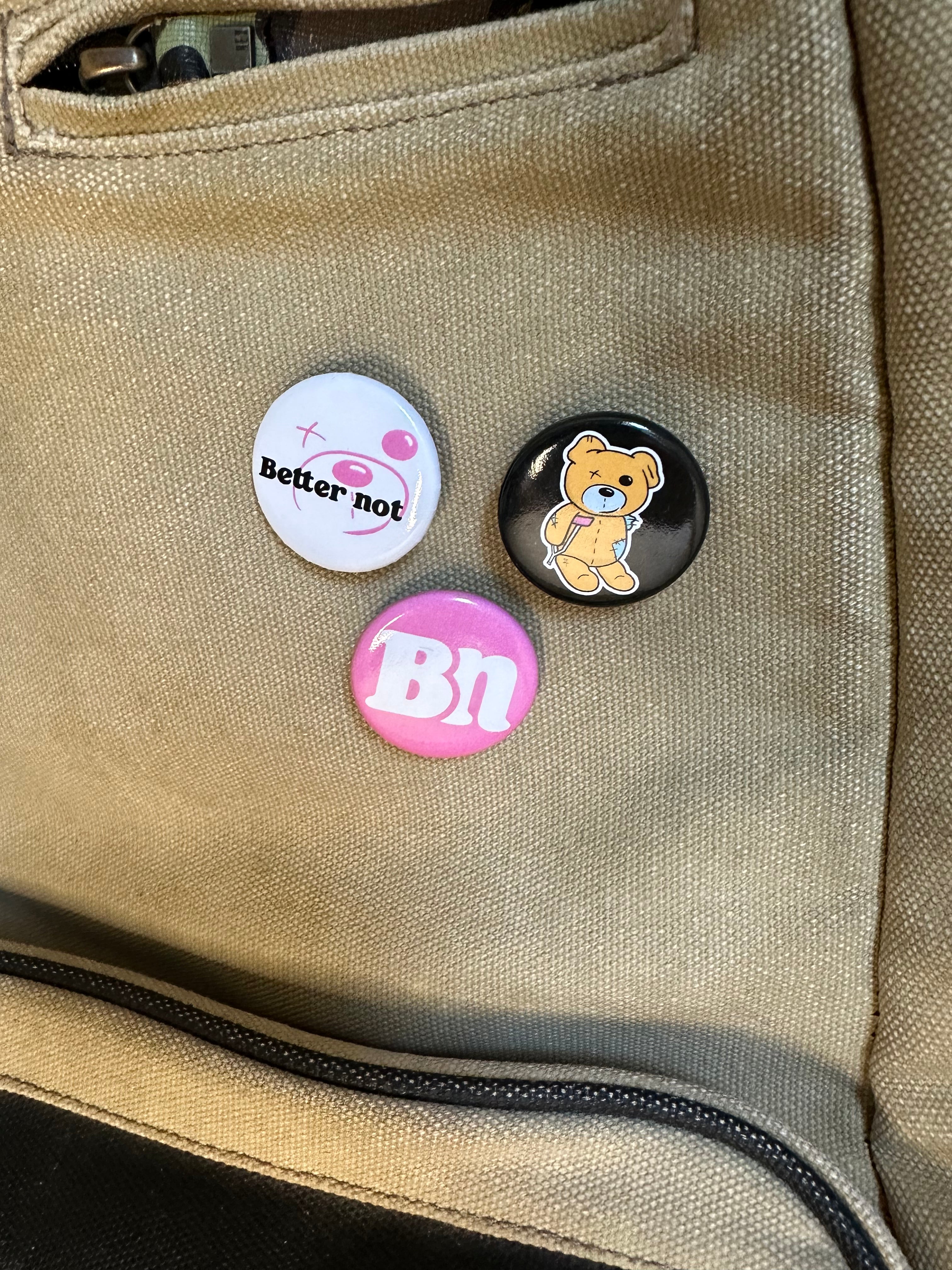 Three 3-Pack Pins on a khaki backpack: white pin with "Better not" and a pink face, black pin with a cute teddy bear, and a pink pin with "bn