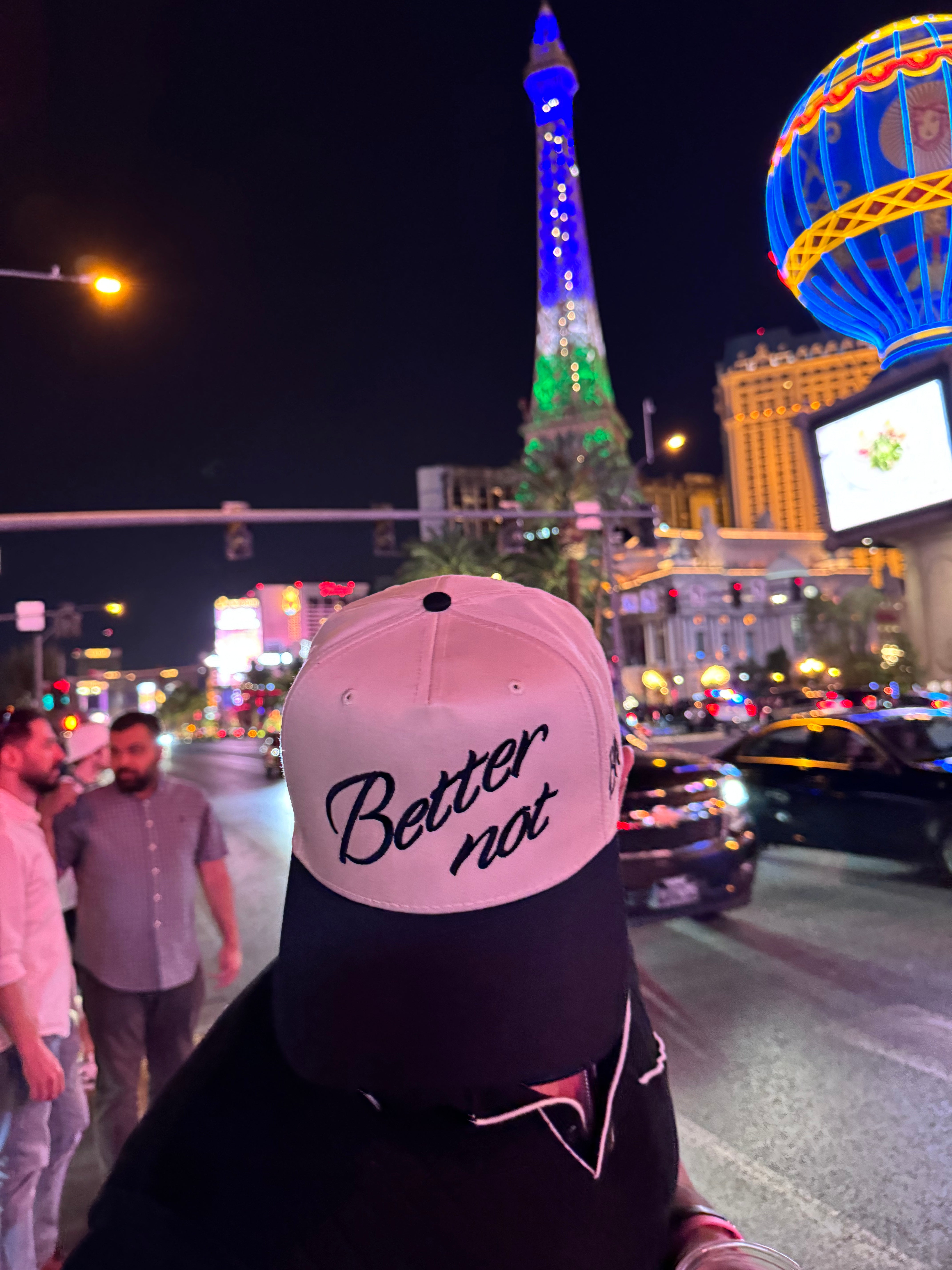A person wearing The Classic by Better not with the phrase "Better not" embroidered on it stands in front of a replica of the Eiffel Tower and neon signs in a bustling city at night.