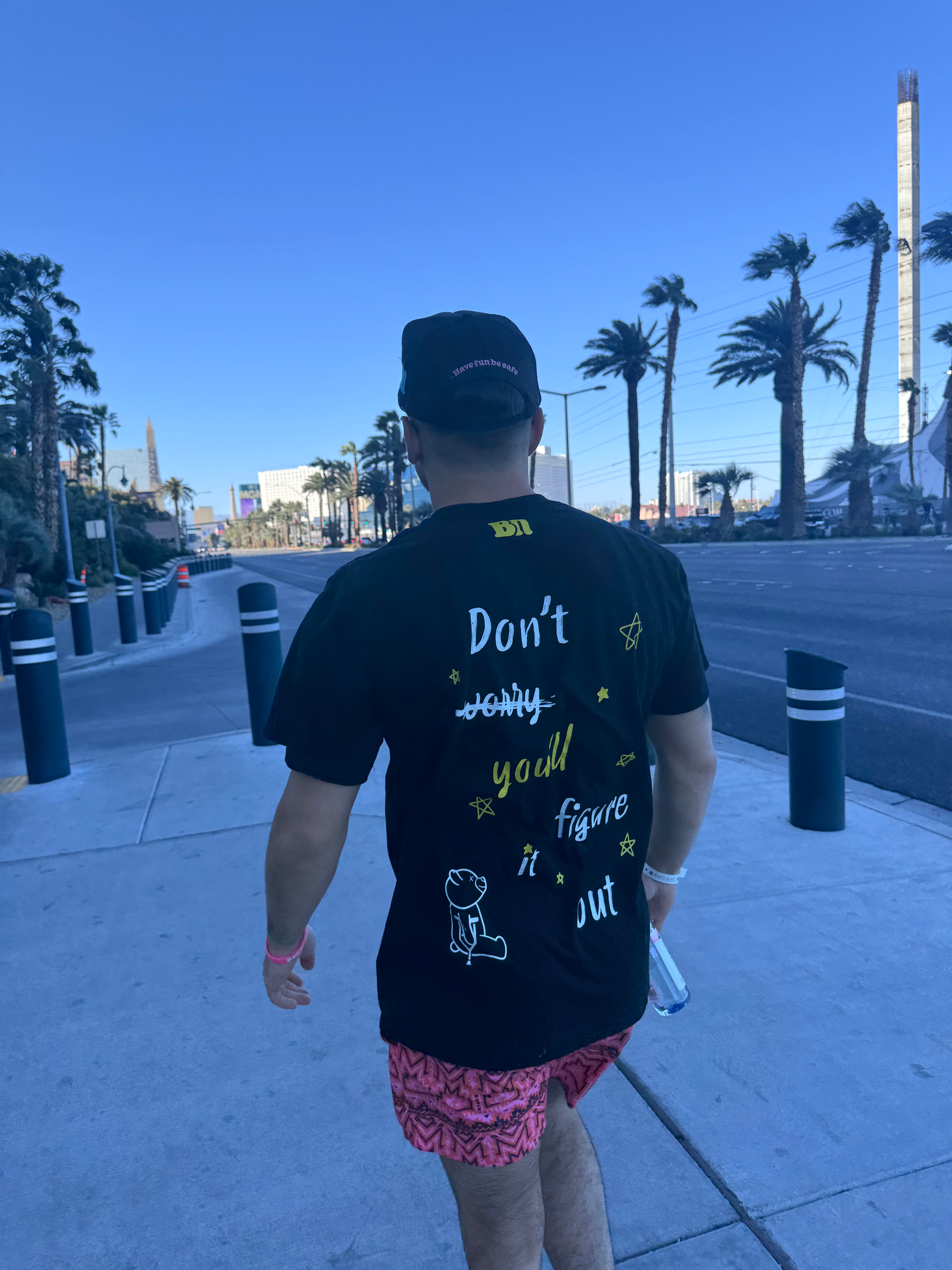 A person wearing a black hat and a black cotton Better not Don't Worry Tee with the text "Don't worry, you'll figure it out" walks down a street lined with palm trees.