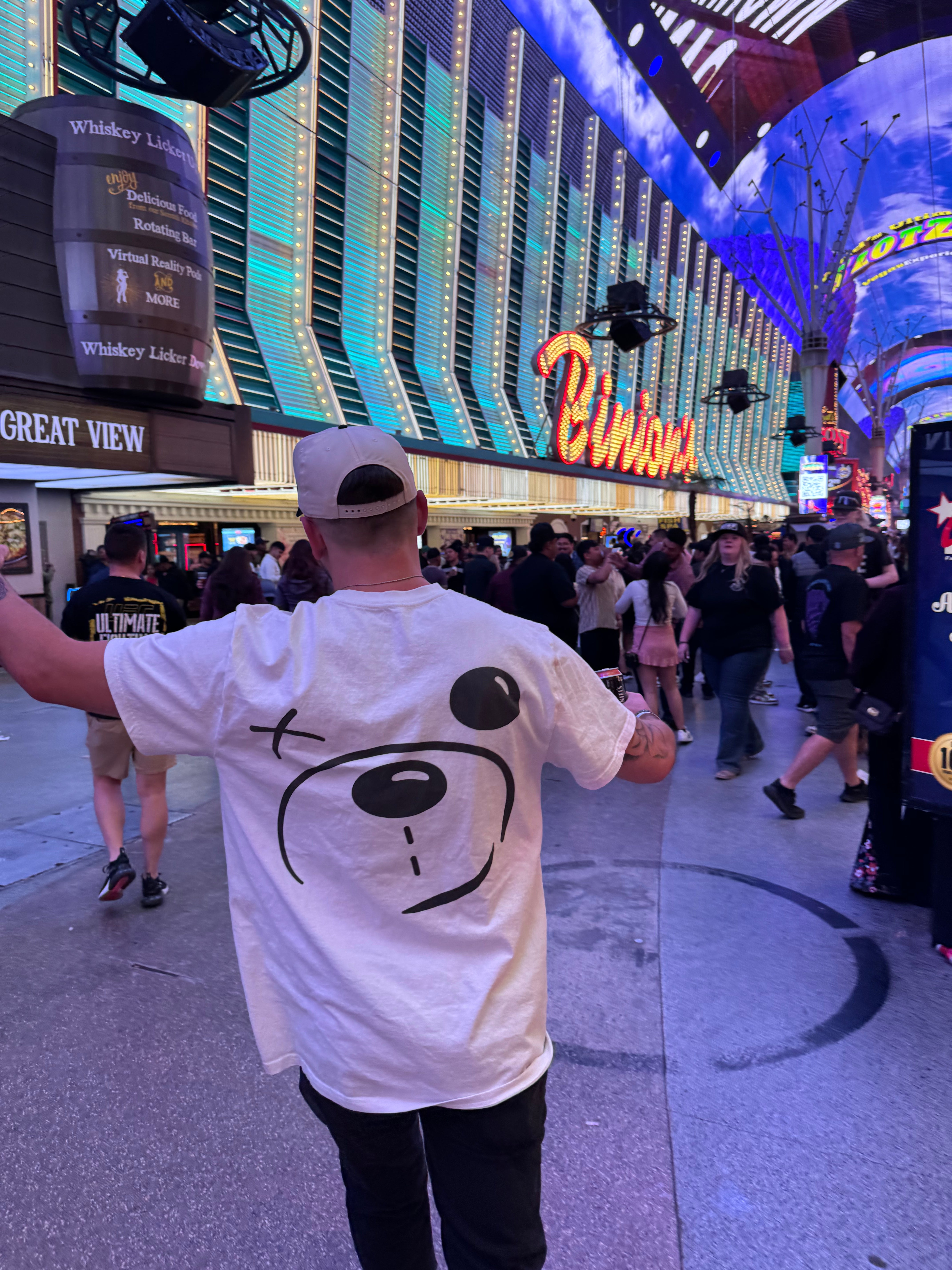 A person wearing a white **Better not Bear Face Tee** raises one arm while walking on a busy, illuminated street in the city. The 100% cotton, unisex shirt stands out amid the bustling crowd and various signs visible in the background.