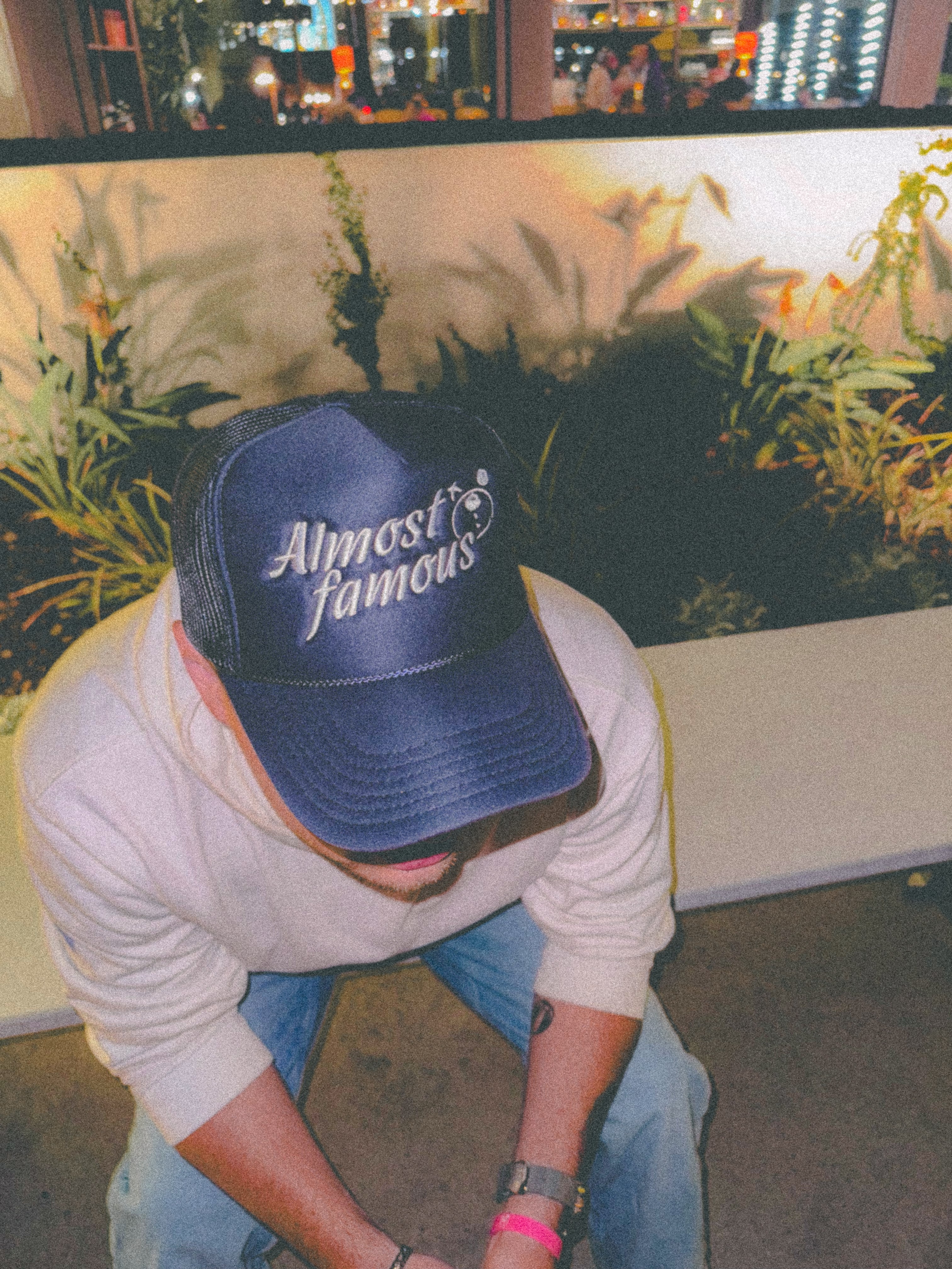 A person wearing an Almost Famous Trucker by Better not sits and looks down, surrounded by plants and city lights in the background.