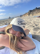 A person wearing a beige and black "Better not The Classic" cap with 3D embroidered artwork is laying on a beach towel, resting their head on their crossed arms. The background features a sandy beach, a few people, and cliffs under a blue sky. This stylish cap also includes snapback closure for the perfect fit.