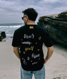 A person wearing a slightly oversized black Better not Don't Worry Tee with the text "Don't you'll figure it out" stands on a beach facing away from the camera. The sky is overcast, and there are large rocks in the background. The unisex cotton shirt adds a touch of casual simplicity to the scene.