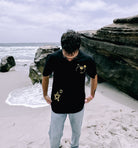 A person wearing a slightly oversized, black unisex cotton Don't Worry Tee by Better not with star designs stands on a beach near large rocks, looking down. The sky is cloudy, and waves are visible in the background.