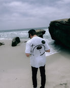 A person in an oversized white Better not Bear Face Tee stands on a sandy beach, facing the ocean, with large rocks nearby.