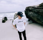 A person wearing an oversized unisex Bear Face Tee by Better not and a black cap stands on a beach near large rocks. The ocean waves and cloudy sky are visible in the background.