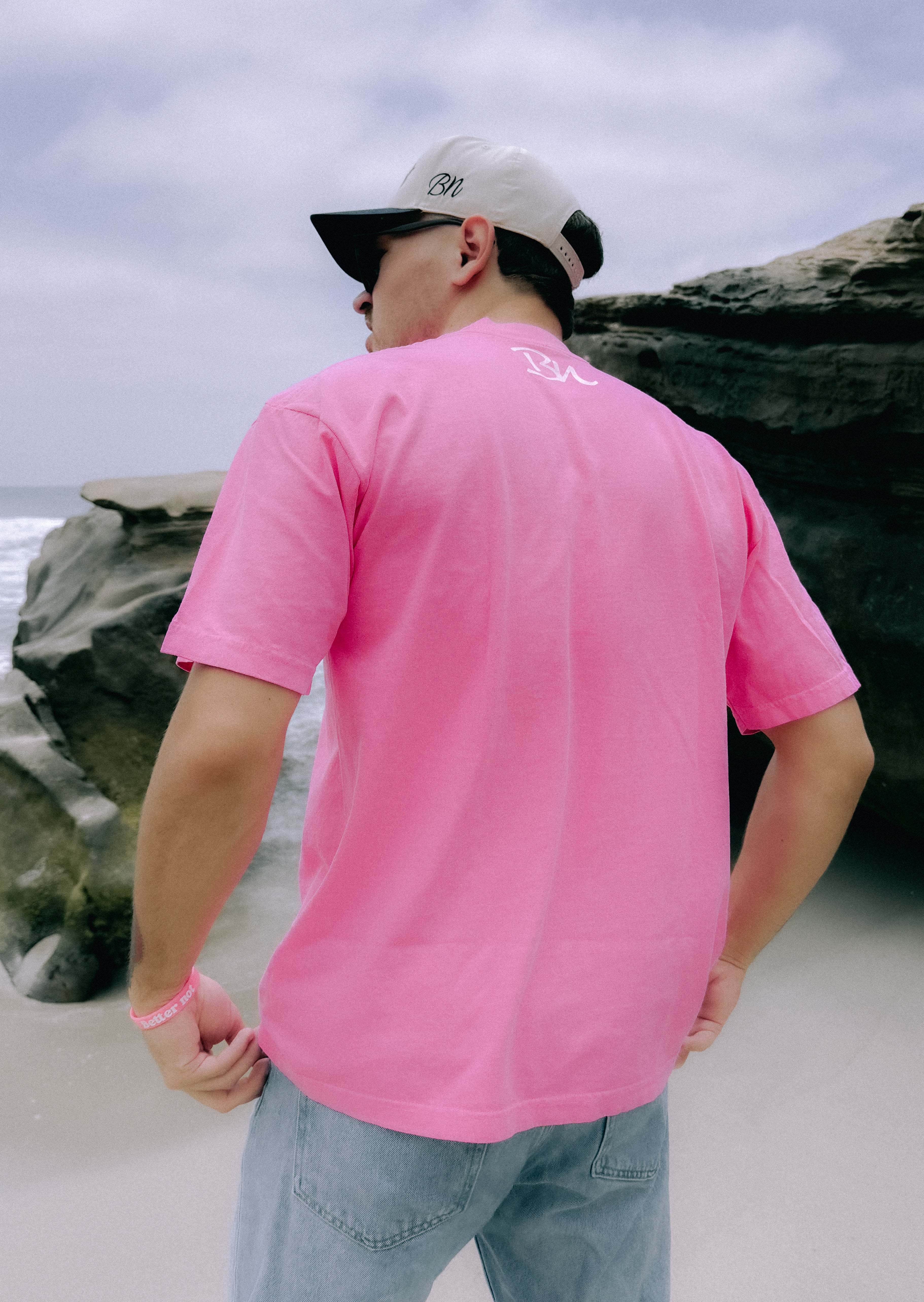 A person facing away, wearing the Off-Centered Tee by Better not made of 100% cotton in pink, paired with light blue jeans, a beige cap, and sunglasses, stands on a beach near large rock formations.