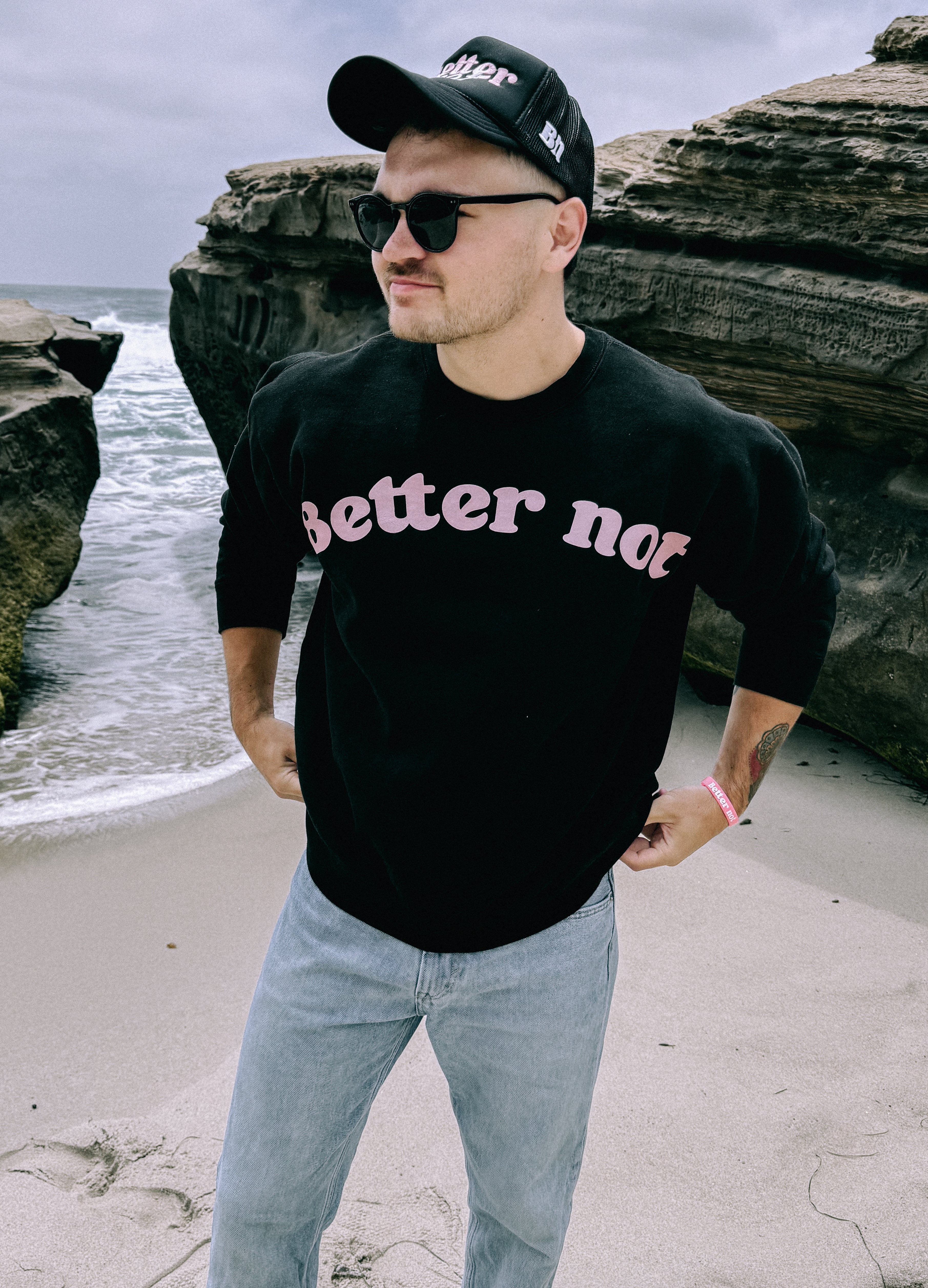 A man stands on a beach, wearing a black "Better not" Staple Crew made of premium ring-spun cotton, sunglasses, and a cap. Rock formations and ocean waves are in the background.