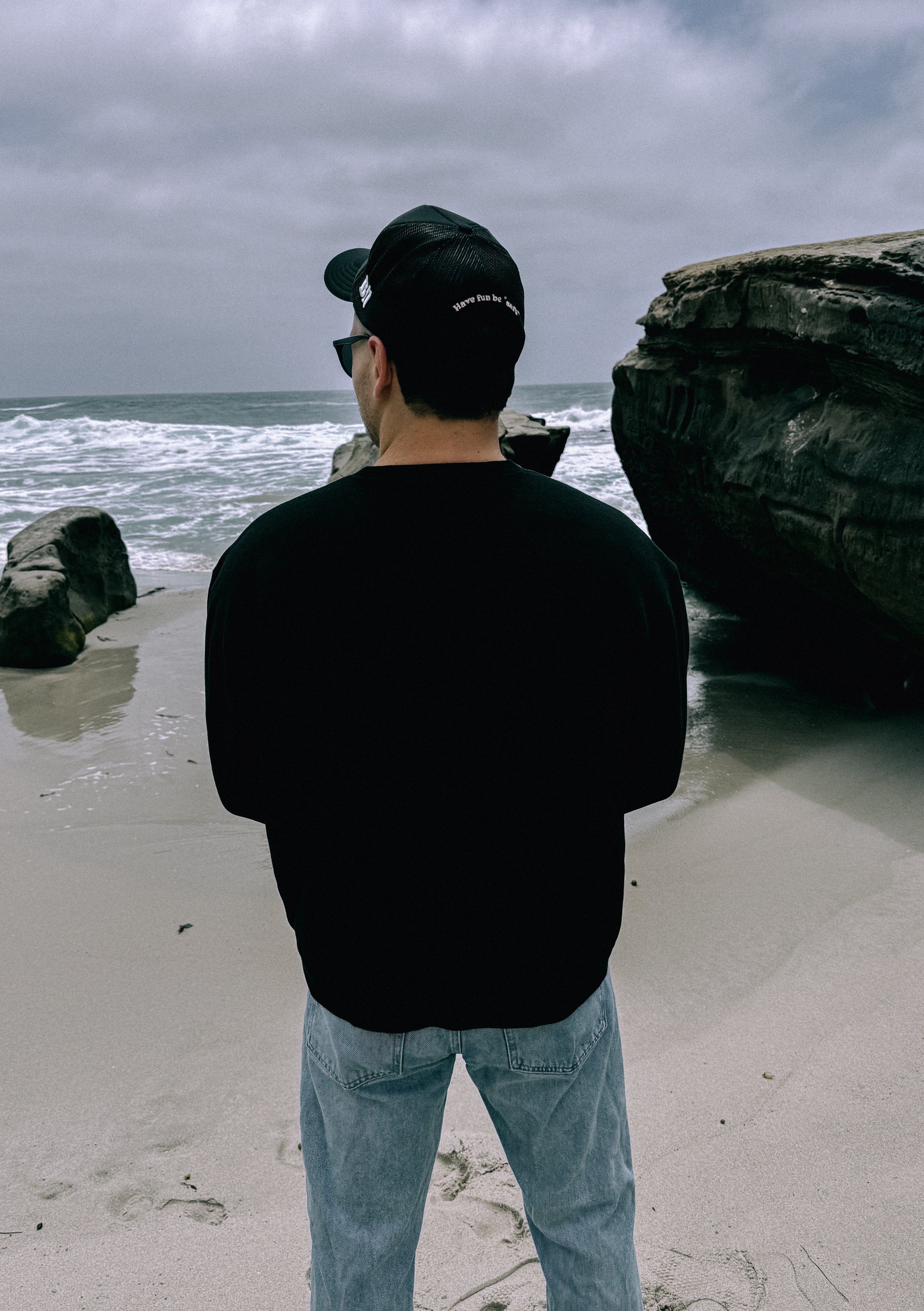 A person in a black Better not Staple Crew, jeans, and a black cap stands on a sandy beach, facing the ocean with large rocks nearby under a cloudy sky.