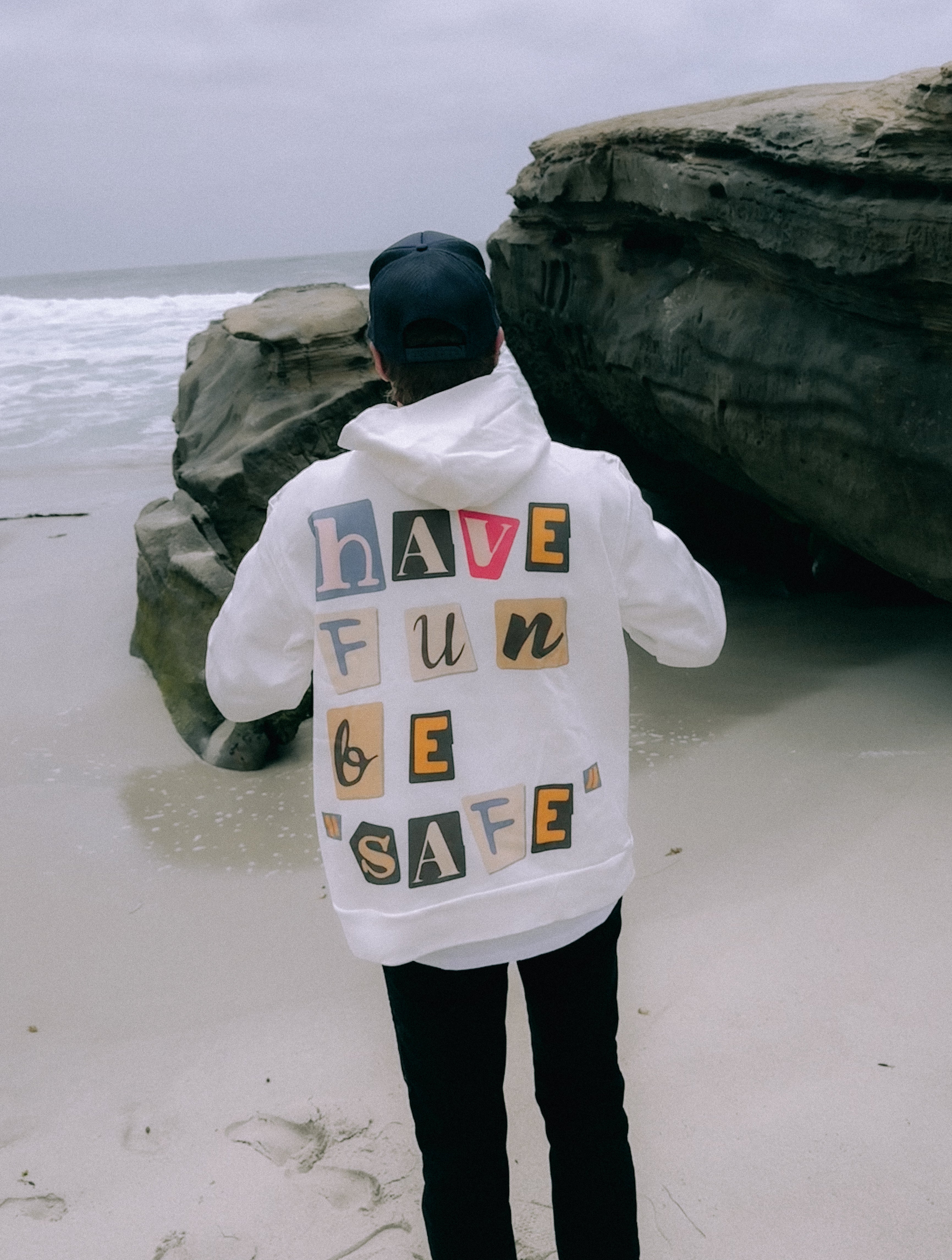 Person wearing a white jacket made from 100% ORGANIC COTTON with "HAVE FUN BE SAFE" written on the back, standing on a beach near large rocks. The person is actually sporting the Have Fun Be "Safe" Hoodie from Better not.