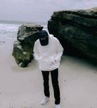 A person wearing a Better not Have Fun Be "Safe" Hoodie, black pants, and a cap is standing on a sandy beach near large rocks with their head down. The ocean is visible in the background.