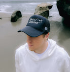 A person wearing a black Better Not Almost Famous Trucker with "Almost Famous" embroidered placements and a white hoodie stands on a beach with large rocks and the ocean in the background.