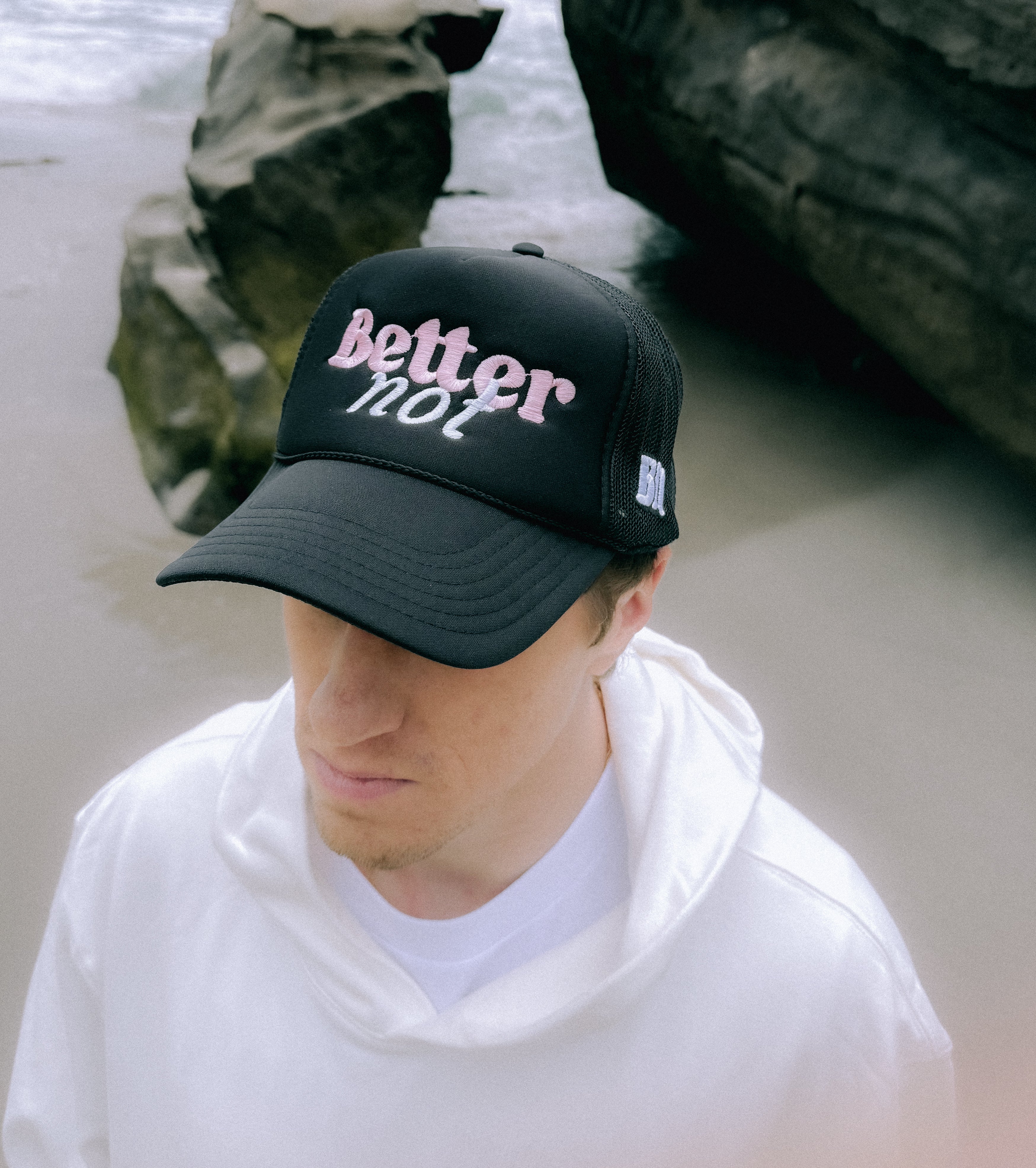 A person in a white hoodie and black cap with "Better not" written on it, designed with a Classic Trucker (mid crown) fit, stands on a beach near large rocks.